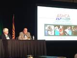 Two anel speakers at the ASHCA conference  with oresentation screen in background