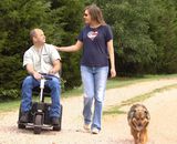 John Enns riding scooter with wife walking next to him and dog in foreground
