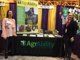 Kelsey on the right with Kaitlyn Higgins on the left in front of AgrAbility table and display with a banner and materials on the table.
