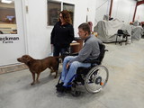 Eric Beckman sits in wheelchair inside farm shop with Emily Freudenburg standing next to him with a dog in between them.