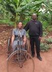 Mutumba Faisal stands next to African woman in wheelchair/bicycle with palm trees in background