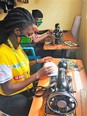 Two african women in a room wearing facemasks like they are making on tredal-style sewing machines