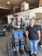 Picture of Kane on the left standing in his tracked stander-wheelchair with Rachel on the right side of the picture next to him inside a large farm implement shed.