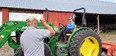 Person in blue sitting on a green John Deere tractor talking to a man with his back to the camera while in the background is a large barn or farm shed that is red with silver metal roof