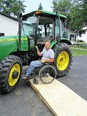 Photo of a man - Doug VerHoeven - in a wheelchair on a ramp that puts him close to a John Deere tractor door - there is a white house and shed in the background