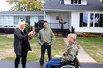 Two people standing in front of a white house with a black porch and hammock on the porch recording a video of older gentleman in a wheelchair.