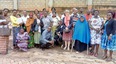 Picture showing a group of African men and women with most of them standing but a few squatting and one in a wheelchair standing on a cement courtyard with a cement block wall behind them.