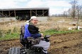 Smiling man on caterpillar-tracked wheelchair on dirt fieeld with open-faced metal pole-barn behind him.