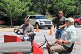 3 men in parking lot 1 standing and holding water bottle with other 2 seated on mock tractor seats with red box in front of them & steering wheel & lever