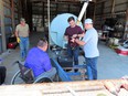 4 men in pole barn working with battery and cables in front of large light-blue water tank. 1 man in lower-left is in standard wheelchair