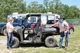 Sideways gray Ranger UTV with woman on left at hood and man at back-both wearing blue jeans & white T-shirts with a blue and a red pickup truck and a camping trailer in background