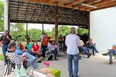 Karen Funkenbusch in blue jeans and pink top with back to camera teaching a group of people seated outside a pole barn under a roof.