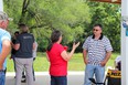 John Fuller in sunglasses - striped gray & white golf shirt & blue jeans speaking with long-brown-haired woman in bright red top & blue jeans with green field and trees in background