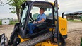 Elderly man in baseball cap-blue t-shirt & bib overalls sitting in cab of tracked bobcat