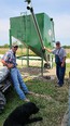 2 men 1 on left and 1 on rt next to a large green bin with silver auger-tube & a black lab dog lying in bottom front of picture