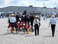 Amish man steering red farm implement with yellow metal wheels drawn by 3 draft horses. Large metal-sided event building in background.