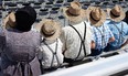 Amish family sitting on bleachers backs to camera Mom on left in black bonnet and holding youngster with only straw hat visible. 3 boys and dad on rt all wearing straw hats checkered shirts and suspenders