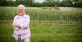 Gray-haired woman with glasses & pink top with arms folded standing in green field with corn growing in background