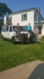 White van in green grass in front of large 2-story white house with green shutters. Ned & Heidi Stoller standing next to van & their 4 children sitting or standing on top of van