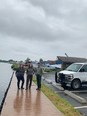 2 women & a man standing on board walk next to body of water with blue & white seaplane behind them & cloudy skies