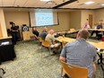 Classroom with Cindy Chastain in black at front talking to people seated at tables in a rectangular formation.