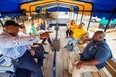 2 African American men in foreground and 5 other people in background all seated on wood benches under yellow & blue canopy top