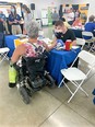 Man in mask behind table with blue table cloth doing health screening of woman in wheelchair in front of him