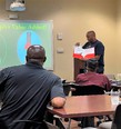 Chef Joseph displaying red folder of materials in front of class with 2 black men in front of him and PowerPoint slide behind him saying VALUE ADDED!