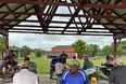 People sitting in a tin-roofed pavilion on a farm with a red barn in the background