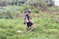 African man in brown baseball cap brown & white t-shirt and dark pants carrying sack in left hand & 3 fish in right while walking through grass near a lake