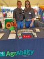 Table in tent with AgrAbility blue table cloth in foreground and 2 women standing behind it in blue jeans and dark gray FarmAgain shirts