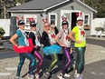 5 women posing with hands on hips in shiny brightly colored fish-scale design spandex pants & red lobster vests over shirts with red lobster claw headbands & gray building with white trim behind them