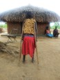 African child on crutches in front of brick hut with grass roof & a man & woman sitting next to hut