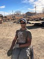 Man in wheelchair on dirt lot wearing gray baseball cap t-shirt pants and gloves with small house & blue sky in background