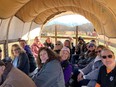 15 people seated in a conestoga wagon with green grass on each side & hills and blue sky in background