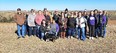 Group picture of 21 people standing on brown grass with prairie in background & black & white dog in front of them