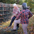 2 women in pants & checkered flannel shirts looking at lobster cages in a fallen-leaf covered lot.