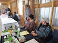 Inside of pole-shed with long tables on left & people behind them on right. Table in foreground has AgrAbility brochures on it and Ed Sheldon in dark vest and flannel shirt seated behind it