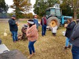 Grass yard with trees and blue Ford tractor in background. 6 people in foreground standing around fired barrels and pots making hog roast