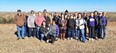 Group picture of 21 people standing on brown grass with prairie in background & black & white dog in front of them