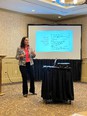 Woman in dark slacks - red blouse - tan jacket teaching at front of room with blue slide on projector screen behind her