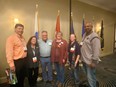 3 men & 3 women standing in front of several flags in a room