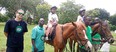 2 horses with an African child one each wearing white t-shirts and riding helmets. 3 Black men holding horses and 1 white man standing to left side of pic