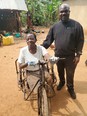 Black African woman in hand-powered wheelchair with black man in black AgrAbility sweatshirt standing on her left and African village scene with banana trees in background.