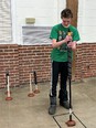 Male youth wearing long black & white jogging pants and green t-shirt standing in room holding a very long-handled toilet plunger vertically in front of him.