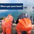 2 people from the back side wearing bright orange chest waders and bending over to get something out of a body of water