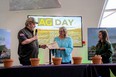 A man with a microphone on the left speaking with 2 women on the right behind a table with flower pots on it and a screen behind all of them that reads AG DAY.