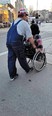A man in a red & white baseball cap and bib overalls pushing a woman in a wheelchair on a city street.
