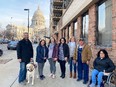 Group of people on city sidewalk with man on left having a guide dog and Madison WI capitol building in background.