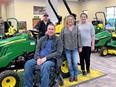 Man in coveralls seated in powered wheelchair in front of John Deere tractor in a showroom with 2 women and a man standing behind him